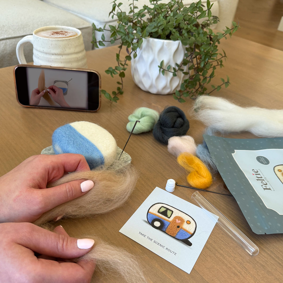 Creating the Happy Camper Feltie using the Feltie Kits tutorial, with wool, needles, tools, and a phone showing the video on a cozy crafting table.