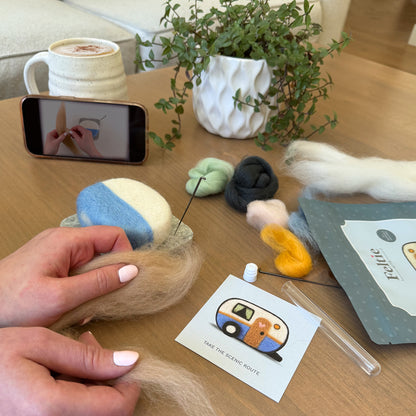 Creating the Happy Camper Feltie using the Feltie Kits tutorial, with wool, needles, tools, and a phone showing the video on a cozy crafting table.