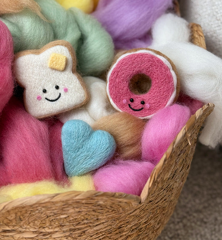 Needle-felted toast, donut, and heart Felties sitting on colorful wool roving from Feltie Kits.