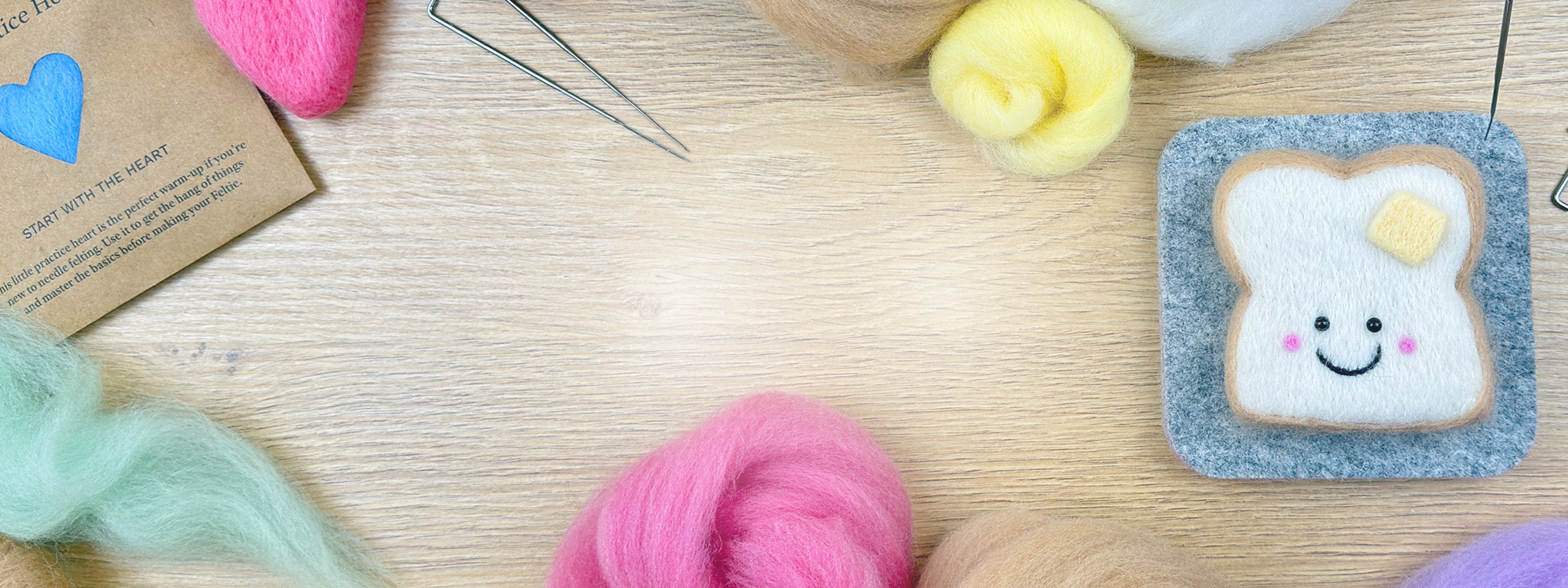 Needle felting kit materials and finished buttered toast Feltie displayed with wool, tools, and the Practice Heart on a wooden table.