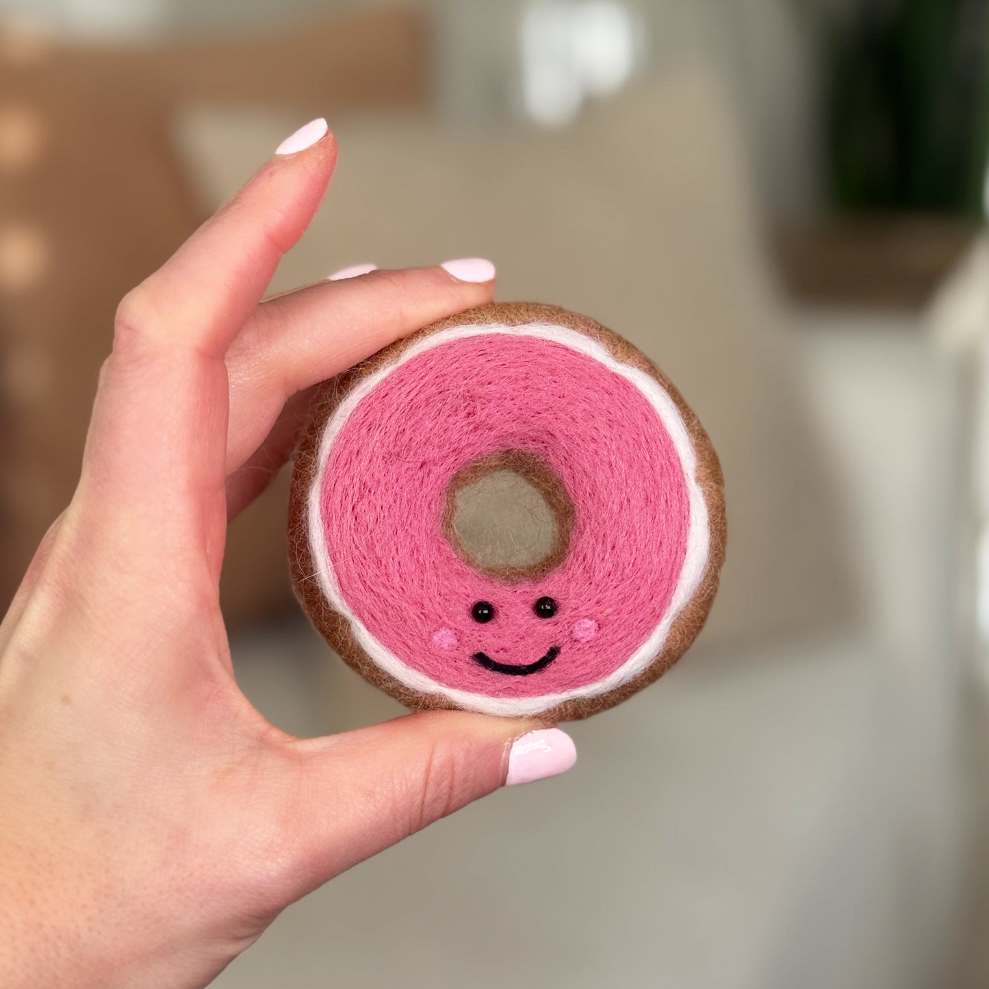 Hand holding the finished Pink Glazed  Feltie from the Feltie Kits needle felting kit, showing the brown donut base, pink icing, white icing and smiling face.