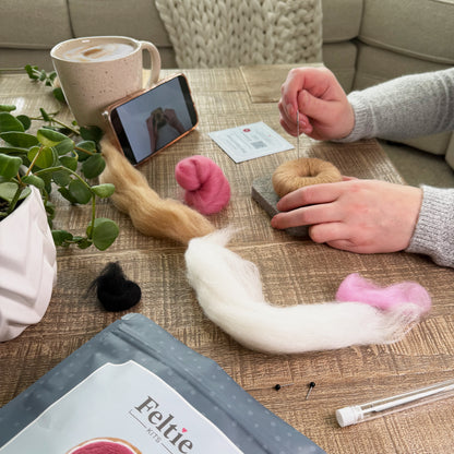 Creating the Pink Glazed Donut Feltie using the Feltie Kits tutorial, with wool, needles, tools, and a phone showing the video on a cozy crafting table.