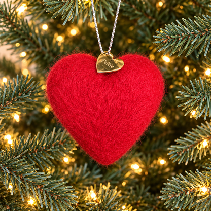 A red handmade needle felted heart Christmas ornament hanging on a Christmas tree, with a gold heart Feltie charm and silver hanging string.