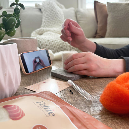 Creating the Salmon Sushi Feltie using the Feltie Kits tutorial, with wool, needles, tools, and a phone showing the video on a cozy crafting table.