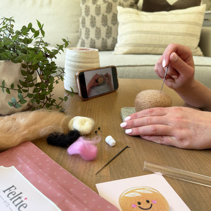 Creating the Sourdough Feltie using the Feltie Kits tutorial, with wool, needles, tools, and a phone showing the video on a cozy crafting table.