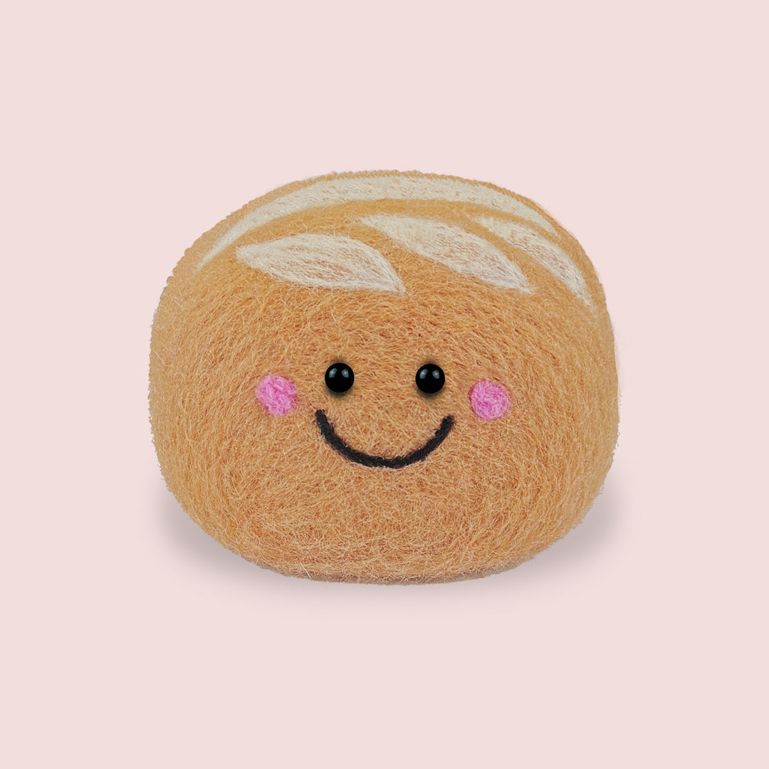 Finished Sourdough Feltie from Feltie Kits, showing a smiling felt sourdough loaf with soft tan wool texture, white scored dough details, and embroidered facial features.