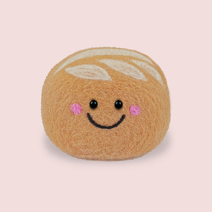 Finished Sourdough Feltie from Feltie Kits, showing a smiling felt sourdough loaf with soft tan wool texture, white scored dough details, and embroidered facial features.