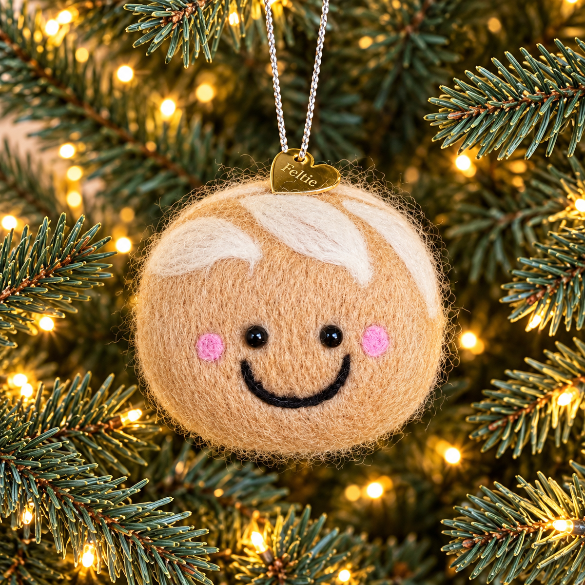 A handmade needle felted sourdough Christmas ornament hanging on a Christmas tree, with a gold heart Feltie charm and silver hanging string.
