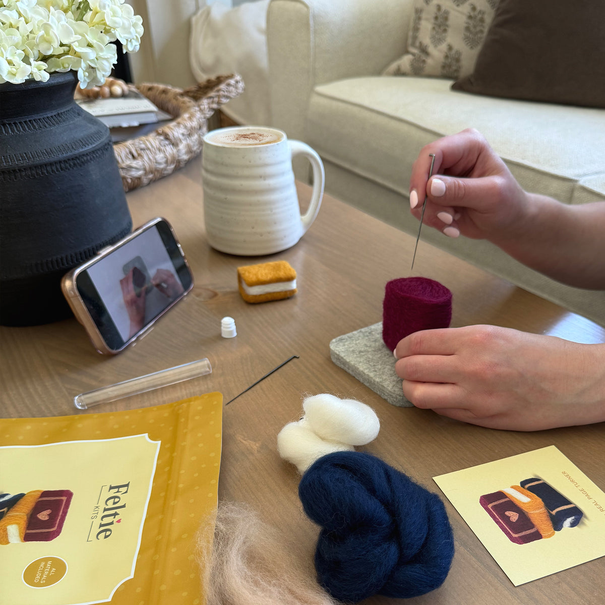 Creating the Storybook Stack Feltie using the Feltie Kits tutorial, with wool, needles, tools, and a phone showing the video on a cozy crafting table.