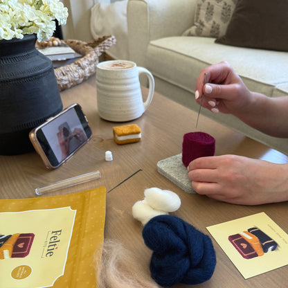 Creating the Storybook Stack Feltie using the Feltie Kits tutorial, with wool, needles, tools, and a phone showing the video on a cozy crafting table.