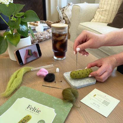 Creating the Big Dill Pickle using the Feltie Kits tutorial, with wool, needles, tools, and a phone showing the video on a cozy crafting table.