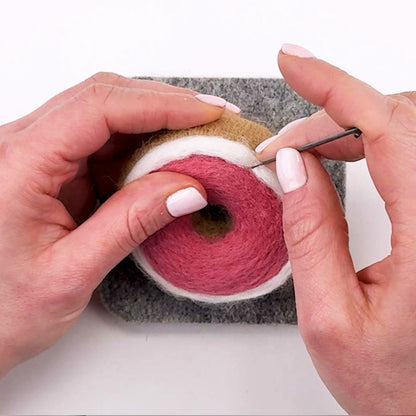 Hands needle felting the  Pink Glazed Donut Feltie from the Feltie Kits tutorial, showing detail work and wool shaping on the felting mat.